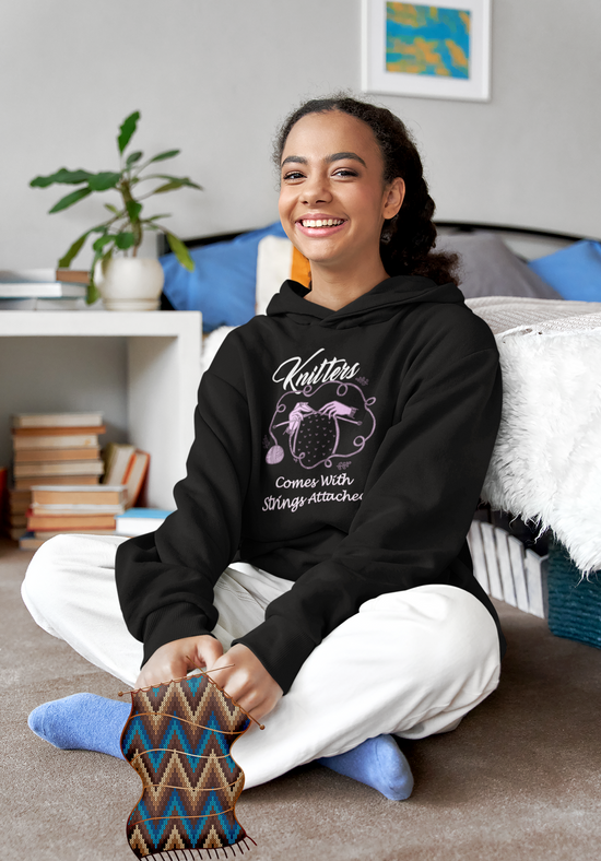 A happy woman sitting on the floor of her bedroom knitting while wearing a black hoodie that says knitters comes with strings attached which is a play on the words.