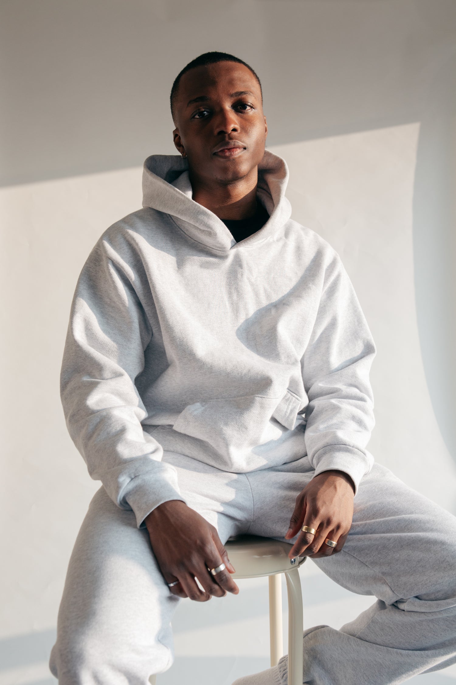 A black male is posing while sitting on a stool. He is wearing a white hoodie with off blue-white trousers.